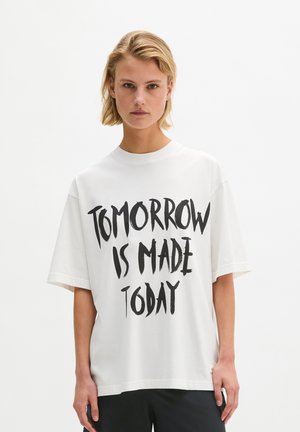 Blond osoba nosząca białą koszulkę z czarnym napisem "Tomorrow is made today", stojąca na jednolitym białym tle.