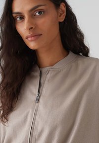 Lichtbeige ritsjack met een gladde textuur, ronde halslijn en minimale stikdetails. Voorzien van een metalen rits en een ontspannen pasvorm.