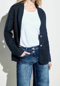 Tmavě modrý lněný blazer s klopovým límcem, dvěma knoflíky a dvěma předními kapsami, spárovaný s bílou košili a modrými džínami.