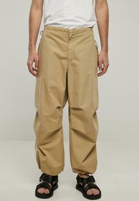 Beige bomuldsbukser med løs pasform, med en front søm, sidelommer og elastisk talje. Sættet er parret med sorte sandaler.