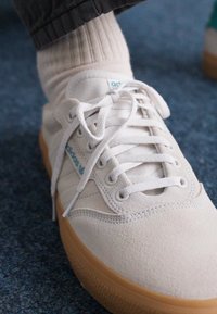 White suede sneakers with a gum sole, featuring round laces, blue Adidas branding, and light grey accents, worn with a cream sock.