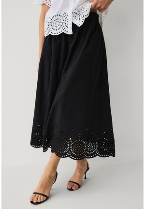 Kobieta nosząca czarną spódnicę midi z kwiatową ażurową lamówką, w połączeniu z białą ażurową bluzką i czarnymi sandałami na obcasie z paskami.