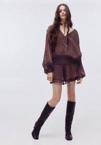 Rjava prosojna bluza z ohlapnimi rokavi, kombinirana z ujemajočo se razširjeno mini krilo in rjavimi usnjenimi škornji do kolen. Teksturiran material, V-izrez.