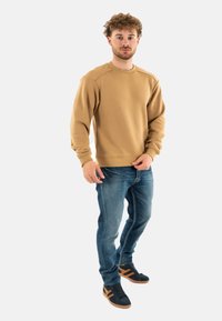 Felpa khaki con collo rotondo, polsini e orlo a costine; abbinata a jeans blu e sneakers nere con dettagli marroni.