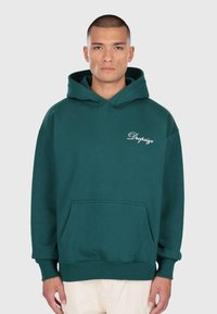 Mann trägt einen dunkelgrünen Hoodie mit Fronttasche und "Dropsize"-Text auf der Brust, steht vor einem einfarbigen hellen Hintergrund.