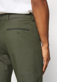 Vue de profil droit d’une personne portant un pantalon vert olive cintré avec une poche arrière à bouton et une chemise blanche rentrée.