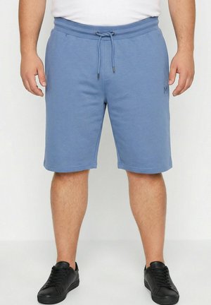 Mann trägt blaue Shorts mit Kordelzug und schwarze Sneakers, steht vor einem einfarbigen hellen Hintergrund.