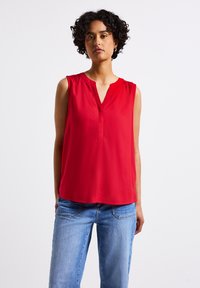Blusa senza maniche rossa con scollo a V, tessuto morbido, pieghe sottili sulle spalle e vestibilità comoda, abbinata a jeans azzurri.