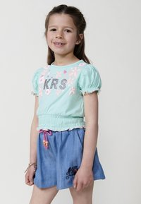 Gaiši zaļš kokvilnas tops ar rozā ziedu izšuvumu un "KRS" ar rhinestones; saskaņots ar ziliem džinsu stila šortiem un rozā aukliņu.