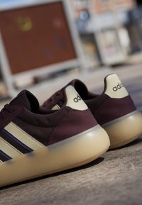 Paire de baskets Adidas violet foncé et crème avec semelles translucides reposant sur une surface en béton à l'extérieur.