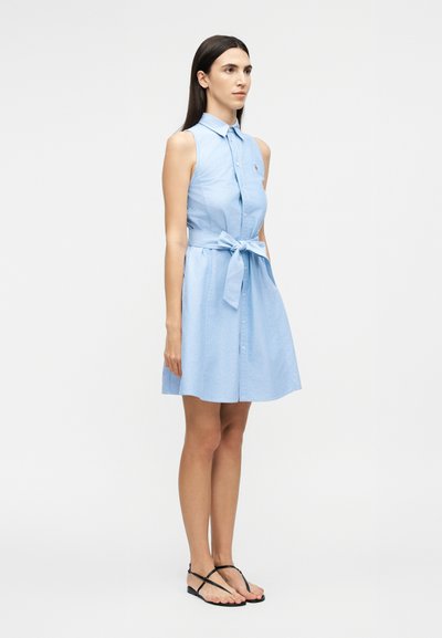 Sieviete stāv, valkājot gaiši zilu kleitu bez piedurknēm ar jostas saiti un melnas sandales ar siksniņām uz vienkārša balta fona.