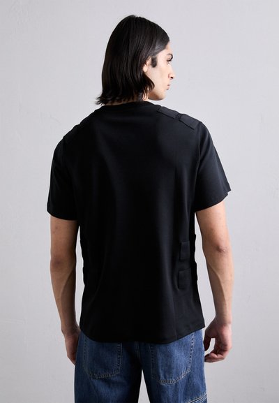 T-shirt nero a maniche corte con una trama liscia, dotato di dettagli hardware regolabili sulle spalle e ai lati.