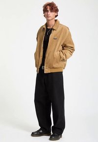 Cordjacke in Hellbraun, mit Reißverschluss vorne, zwei Seitentaschen, schwarzes Logopatch. Kombiniert mit schwarzen, weit geschnittenen Hosen und schwarzen Schuhen.