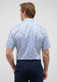 Korte mouwen blouse van lichtblauwe stof met een bladpatroon in donkerdere blauwtinten. De achteraanblik toont een klassieke kraag en een rechte zoom.