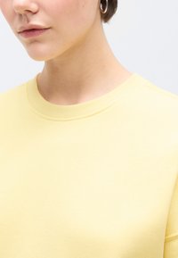 Κίτρινο φούτερ με ριμπ ύφανση στο λαιμό και απαλό ύφασμα. Το κοντινό πλάνο τονίζει τη γραμμή γύρω από το λαιμό και την απαλή υφή του υφάσματος.