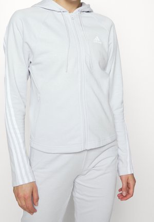Personne portant un sweat à capuche zippé Adidas gris clair avec trois bandes blanches le long des manches et un pantalon assorti, sur un fond blanc.