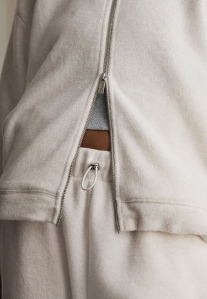 Weiße Fleecejacke teilweise geöffnet, zeigt graues Shirt und weiße Hose mit Kordelzug und Metallverschluss.