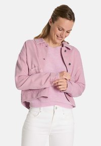 Rosa Denimjacke mit lockerem Schnitt, zwei Fronttaschen und Knopfverschluss, über einem hellrosa Strickpullover getragen. Weiße Jeans runden den Look ab.