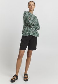 Blusa floreale verde con dettagli bianchi e rosa, dotata di scollatura rotonda e maniche lunghe, abbinata a pantaloni corti neri fino al ginocchio e sandali chunky.