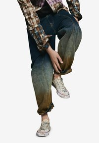 Farmernadrág, amely sötétkéktől világoskékig fokozatosan világosodik a bokánál, kopott, világos színű sportcipőkkel párosítva. Látható egy flanel ing is.