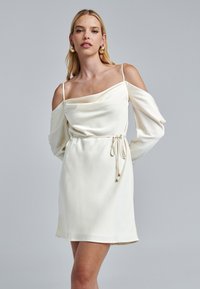 Biela off-shoulder šaty s drapovaným výstrihom, dlhými rukávmi a viazaným pásom. Vyrobené z hladkej tkaniny s jemným leskom.