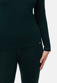Nahaufnahme einer Person, die ein enganliegendes, dunkelgrünes Langarmoberteil und passende Leggings trägt, die Hand ruht am Oberschenkel, mit einem kleinen roten Flaggenetikett am oberen Saum.