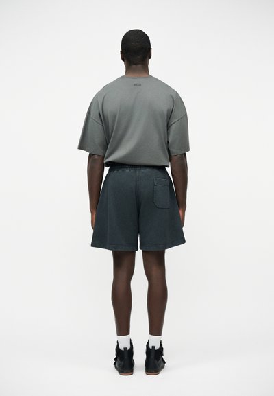 Homme debout de dos portant un T-shirt gris ample, un short sombre avec poche arrière, des chaussettes blanches et des chaussures montantes noires.