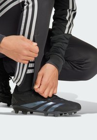 Čierne futbalové kopačky s modrými a striebornými pruhmi, šnurovací dizajn a textúrovaný povrch; osoba sa prispôsobuje zipsu čiernych športových nohavíc.