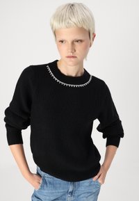 Črni rebrasti pulover z okroglim izrezom, okrašenim z vrsto prozorno svetlečih detaljev. Ima sproščene rokave in ohlapen kroj.