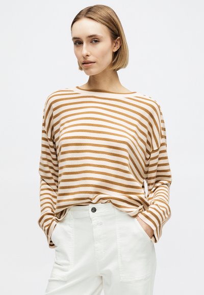 Rövid szőke hajú nő bézs-fehér csíkos hosszú ujjú felsőben és fehér, magasderekú nadrágban, kezét a zsebében tartva.