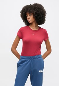 Κόκκινο στενό T-shirt με κοντά μανίκια και στρογγυλή λαιμόκοψη, με μικρό λευκό λογότυπο της Nike. Συνδυάζεται με μπλε φόρμα παντελόνι με κορδόνι στη μέση.