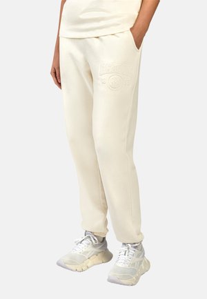 Persoon draagt off-white Reebok sweatpants met elastische boorden en witte chunky sneakers, staand met één hand in de zak.