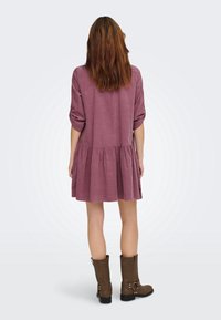 Femme aux cheveux bruns portant une robe mauve mi-longue au genou et des bottes marron mi-mollet, debout de dos, sur un fond clair uni.