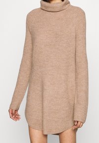 Pull en tricot beige avec un col montant, des manches longues et une coupe légèrement oversize, doté d'un tissu texturé et doux, avec un ourlet arrondi.