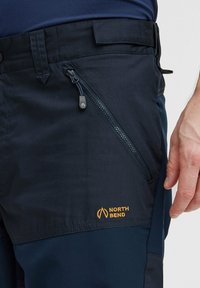Jūras spēku pārgājienu bikses, kas izgatavotas no izturīgas auduma, ir aprīkotas ar sānu ar rāvējslēdzēju slēdzamu kabatu un oranžu "North Bend" logotipu augšējā gurnu daļā.