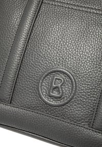 Czarna skórzana torba z teksturowaną powierzchnią, tłoczonym logo w okrągłej ramce oraz szczegółowym przeszyciem wzdłuż krawędzi.