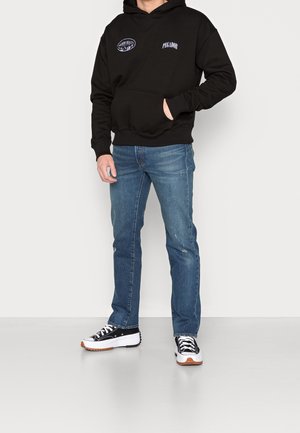 Sudadera con capucha negra con bolsillo frontal y logotipos bordados, combinada con unos vaqueros de mezclilla azul claro y zapatillas negras y blancas.