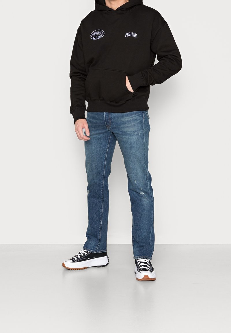 Czarna bluza z kapturem z kieszenią z przodu i haftowanymi logo, zestawiona z jasnoniebieskimi dżinsami i czarno-białymi sneakersami.