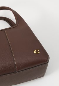 Bolso de cuero marrón texturizado con detalles cosidos y un pequeño emblema dorado "C" sobre un fondo blanco.