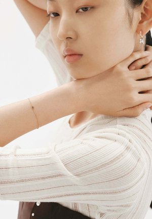 Pulsera de oro rosa con un pequeño charm de cruz en una muñeca descubierta, superpuesta a una blusa blanca texturizada de rayas con cuello redondo.