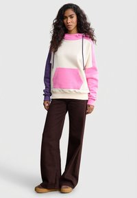 Bluza z kapturem w kolorach kremowym, różowym i fioletowym z przednią kieszenią i kapturem na sznurek, zestawiona z ciemnobrązowymi spodniami o szerokich nogawkach.