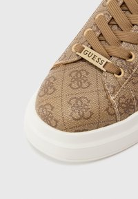 Καφέ πάνινο αθλητικό παπούτσι με υφή μοτίβου μονόγραμμα, διαθέτει λαστιχένια σόλα και χρυσή ετικέτα με την ένδειξη "GUESS".