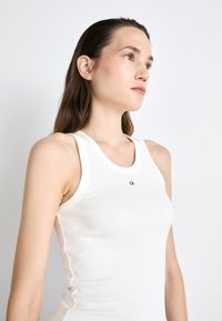 White ribbed tank top with a scoop neck and logo detail in black. Material appears soft and fitted, with wide shoulder straps and no embellishments.