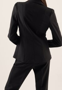 Schwarze taillierte Blazerjacke mit figurbetonter Silhouette, Reverskragen und strukturierten Nähten. Getragen mit passenden schwarzen Hosen. Glatte Textur des Stoffes.