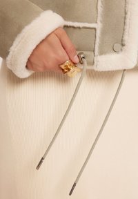 Zielona zamszowa kurtka z białym futerkiem; ręka trzyma regulowany sznurek z srebrnymi akcentami; złoty pierścionek na palcu; kremowa spódnica w prążki.