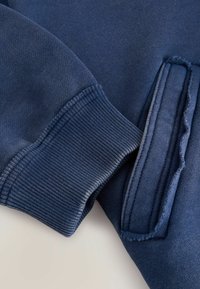 Mikina navy modré barvy s vroubkovanými manžetami a vnitřní kapsou. Hladký povrch s viditelnými švy. Jednoduchý, funkční design.