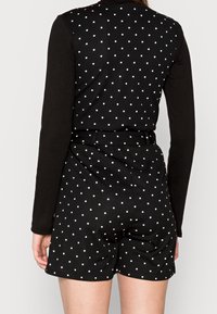 Svart långärmad bodysuit med prickigt mönster på shorts-delen; har en åtsittande design och ett cinch-bälte i midjan.