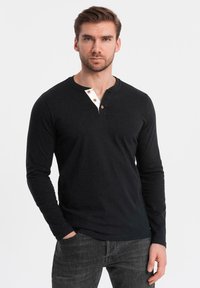 Schwarzes langärmliges Henley-Shirt mit einer weißen Blende und drei Knöpfen. Weicher Baumwollstoff mit figurbetonter Passform. Lässiges Design.