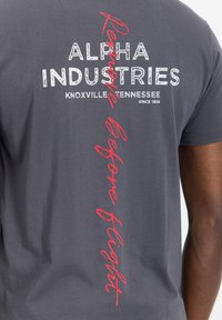 Šedé bavlněné tričko s výstižným bílým textem "ALPHA INDUSTRIES, KNOXVILLE TENNESSEE" a červeným kurzívním textem "Remove Before Flight" na zadní straně.