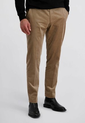 Man die beige corduroy broek draagt met zijzakken en zwarte leren Chelsea boots, staand tegen een effen lichte achtergrond.